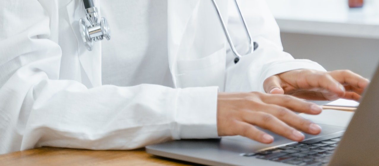 A Woman in White Lab Coat Smiling while Typing on Laptop | Breast Cancer Car Donations