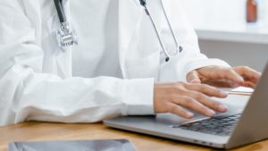 A Woman in White Lab Coat Smiling while Typing on Laptop | Breast Cancer Car Donations