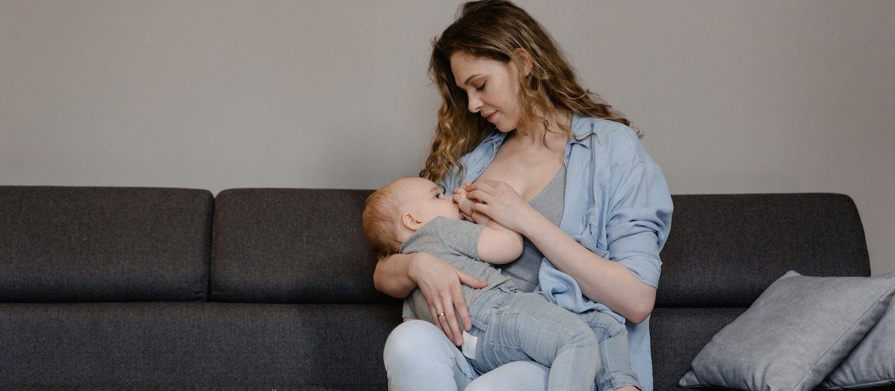A Woman Breastfeeding Her Baby while Sitting on the Couch | Breast Cancer Car Donations