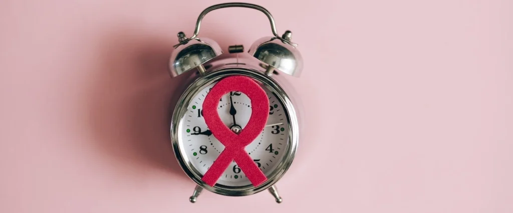 Silver alarm clock with pink breast cancer awareness ribbon on a pastel pink surface, symbolizing support