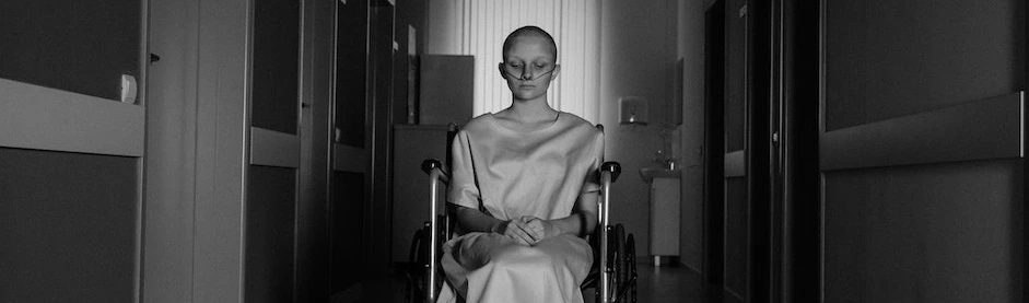 A woman in a hospital gown sits in a wheelchair in a dimly-lit hallway, evoking a somber mood