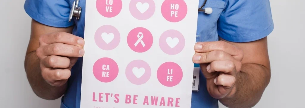 Smiling doctor in blue scrubs holding a breast cancer awareness poster with pink symbols